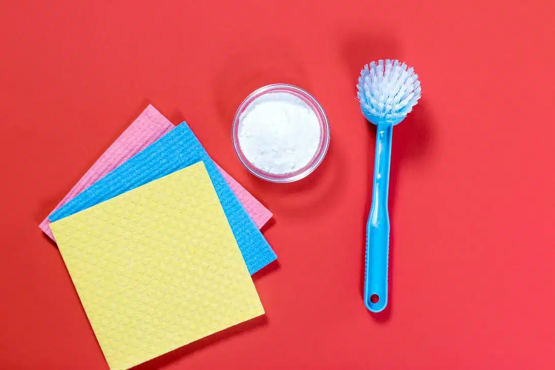 blue scrubbing brush next to baking soda and 3 microfibre cloths on a red background