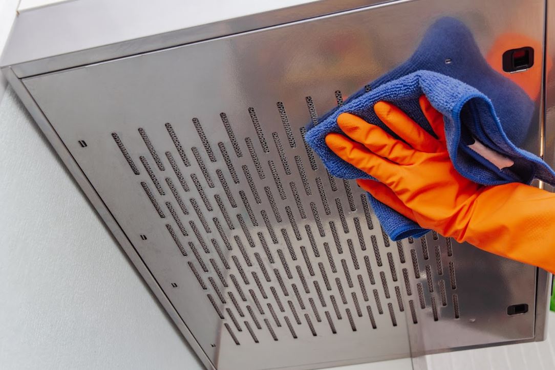 person wearing orange gloves using a blue microfibre cloth to clean rangehood and filter